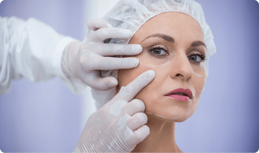 Woman with a cap on. Her face is being examined for dermal fillers by two gloved hands.